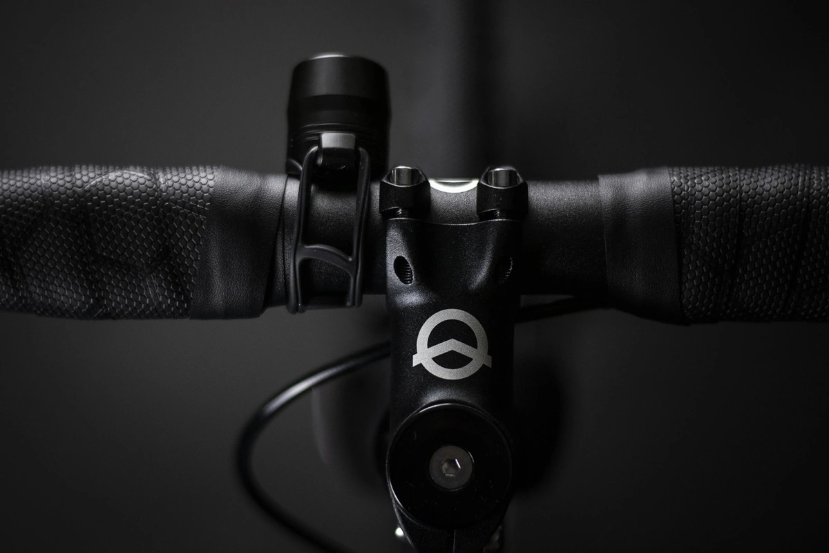 Top view of a handlebar with a Reflection Mini front light on its silicone strap to the left of the stem, black bar tape and a stem with a round logo