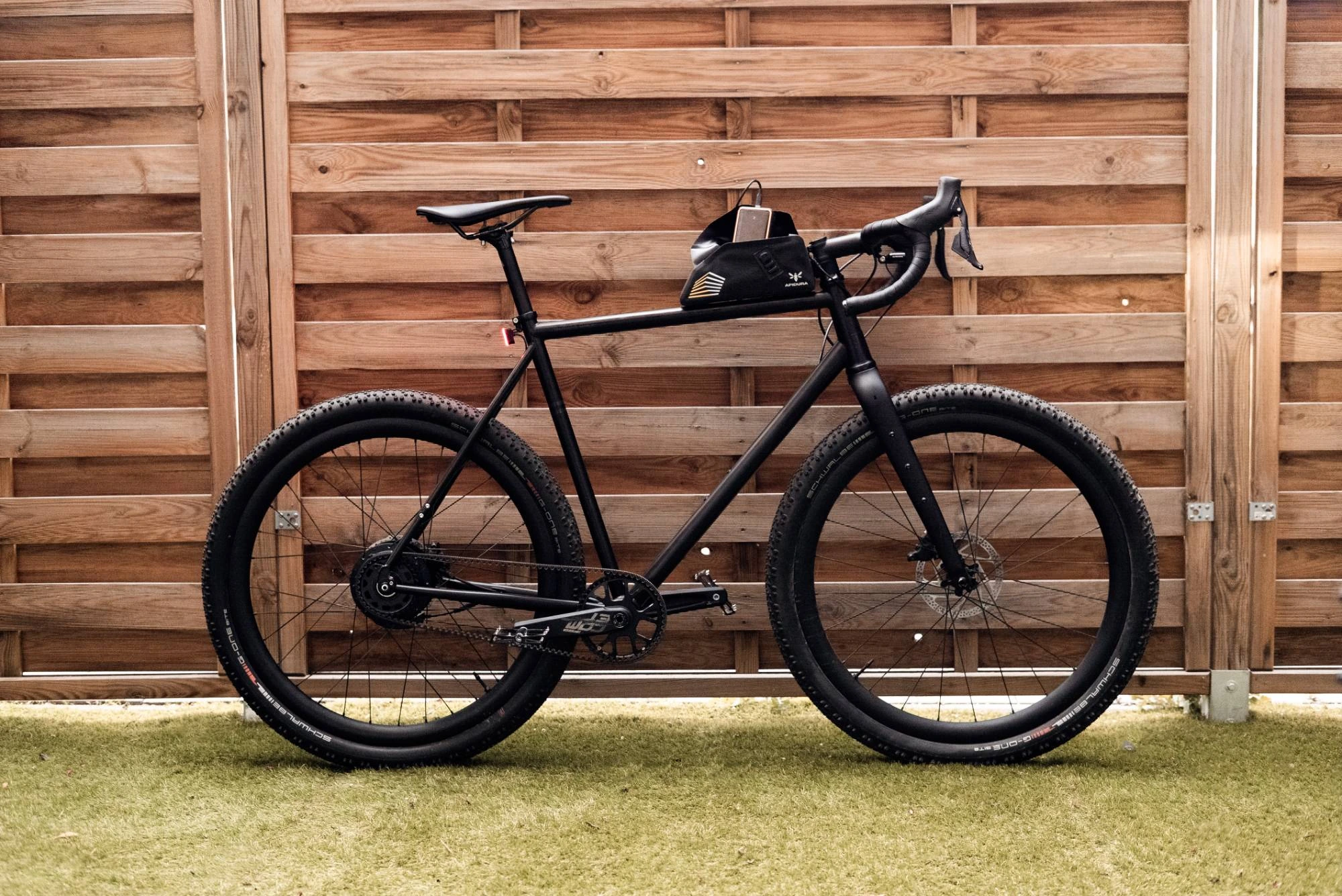 Booda Bike Alpine 3x3 gravel bike in side view in front of a wooden fence, with the Apidura top tube bag and a power bank, belt drive and bent handlebar