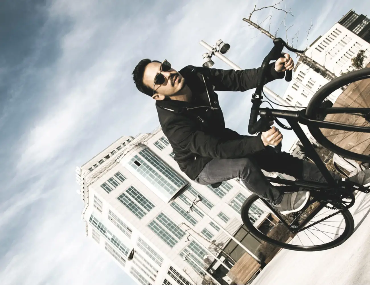 A rider in sunglasses leans into a turn on a Booda Bike next to a modern building