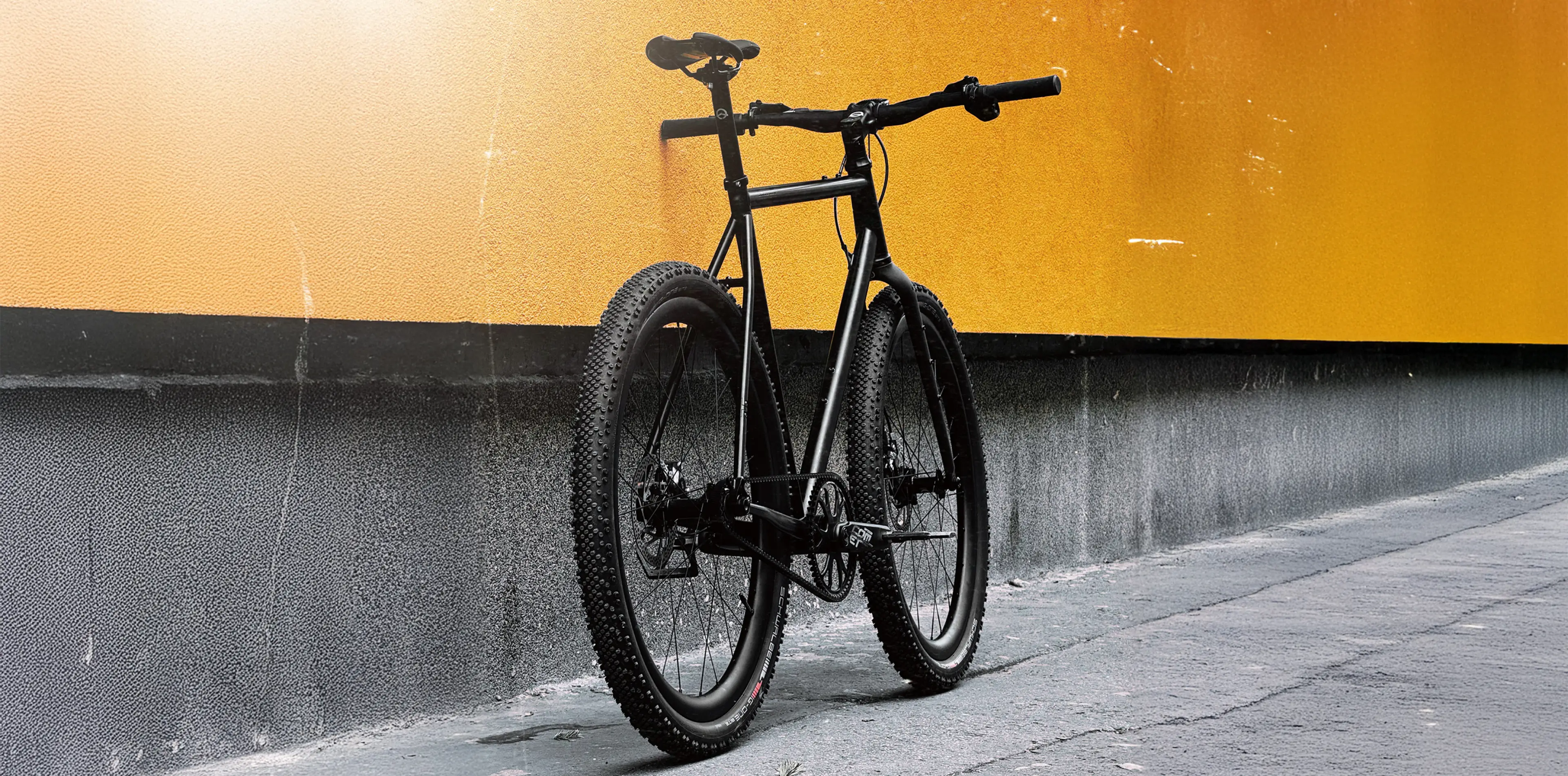Black Booda Bike Sander single-speed gravel bike with a straight handlebar, standing next to an orange wall
