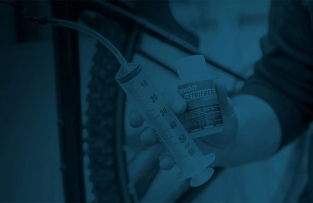 Hand holding a Pinion gearbox oil bottle and an oil syringe with a clear hose during an oil change, in blue tone